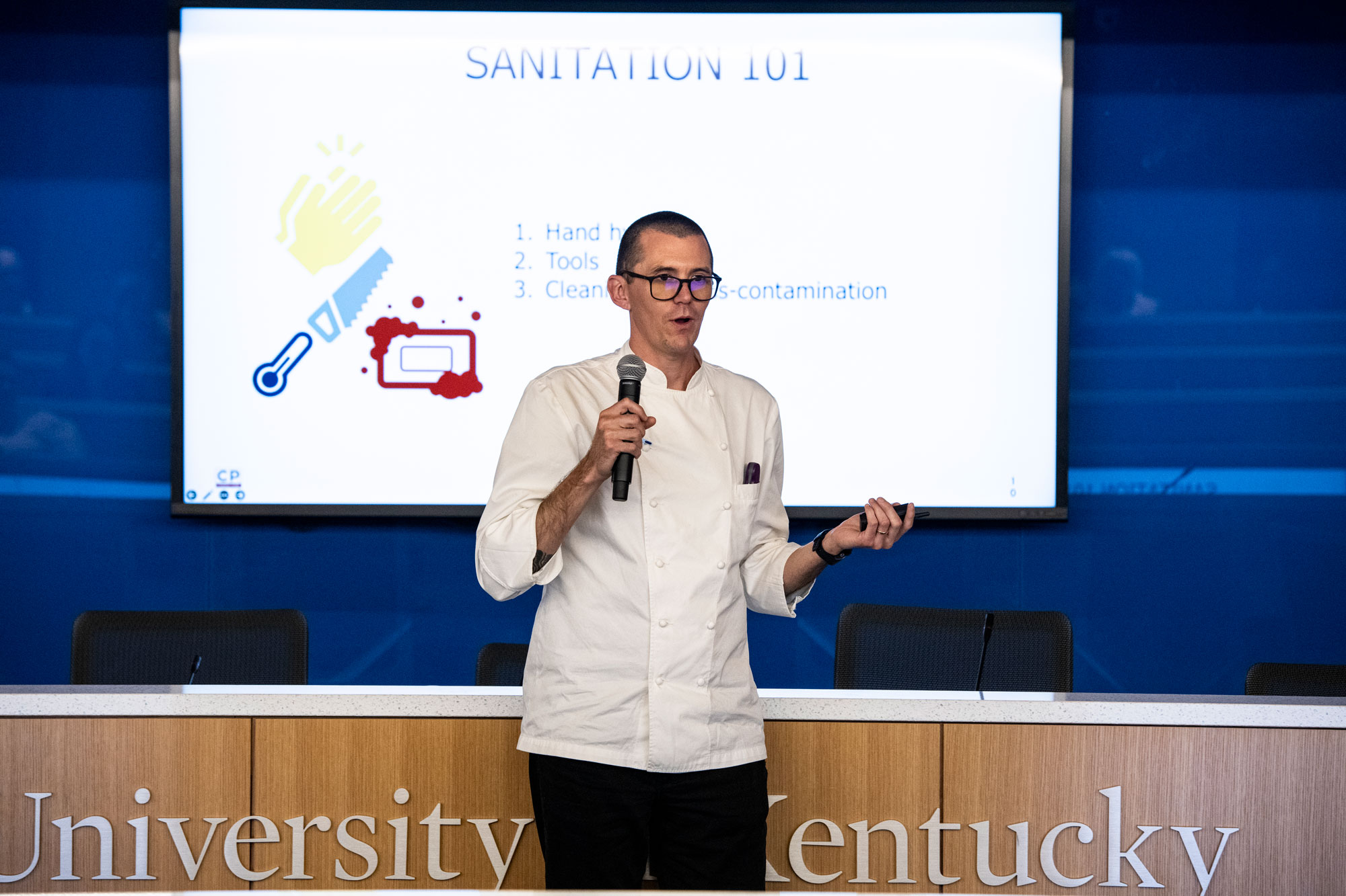 Trevor, a tall, thin white man with glasses wearing a lab coat standing in front of a slide projection that reads "Sanitaiton 101"